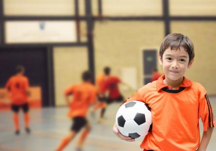 A young child holds a soccer ball while his teammates play a game in the background