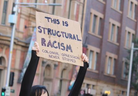 Image of a handmade sign at a protest, with the words 'This is structural racism. Colonial oppression never stopped'