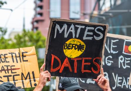 Depicts hand-made signs at a public demonstration, focussing on a sign that says 'Mates before Dates' over a painted Aboriginal Flag