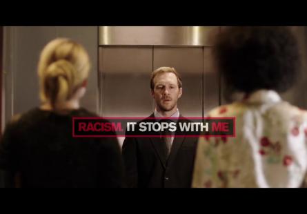 Depicts two people looking into an elevator, with a man on the inside of the elevator standing and looking back out. Overlaid is the Racism. It Stops With Me logo, depicting the words 'Racism. It Stops With Me' in red and white on a black background