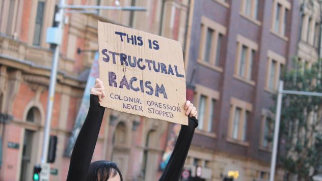 Person holding up sign at anti-racism protest. Sign reading 'This is structural racism. Colonial oppression never stopped'.