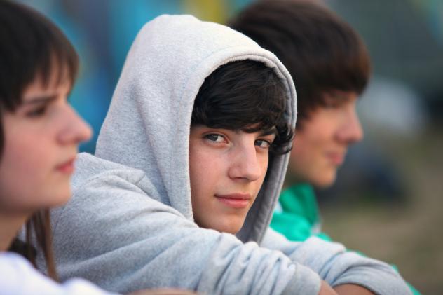 Three teenagers sitting next to each other. Boy in the middle is wearing a grey hooded jacket and is looking directly to camera.